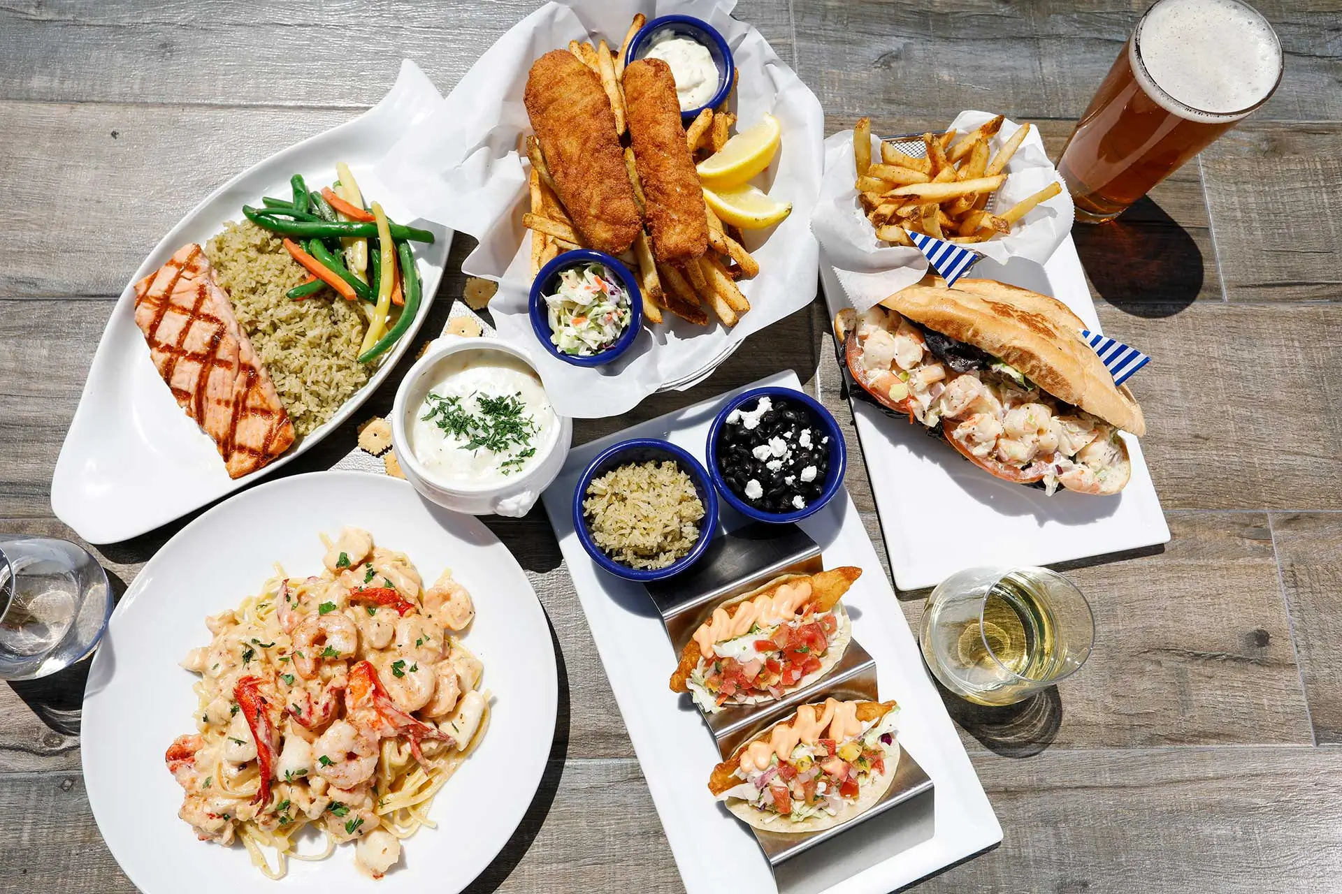Overhead view of a seafood spread at Seaside on the Pier with fish and chips, lobster roll, grilled salmon, shrimp pasta, fish tacos, and fries on a wooden table.