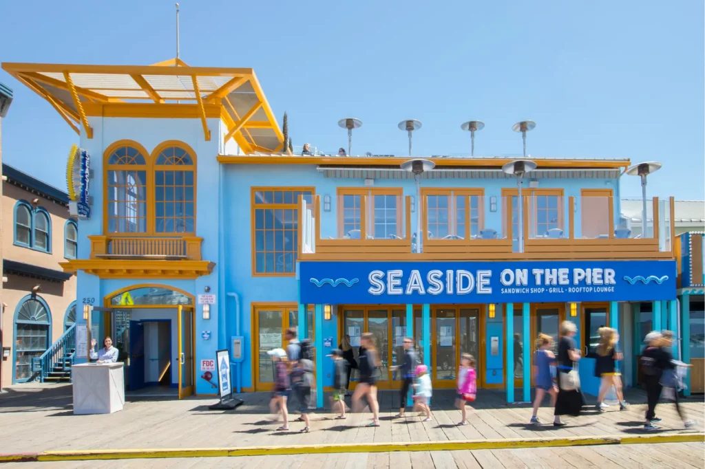 Seaside on the Pier exterior on a sunny day, bright blue building with yellow trim, rooftop lounge, and pedestrians walking along the wooden pier in front.